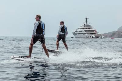 Two men on jetboards race across the water with a luxurious yacht anchored in the background.