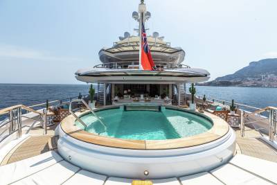 A luxurious yacht features a stunning circular pool on its deck, overlooking the sparkling blue ocean and a distant, picturesque coastline.
