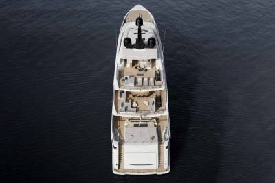 An aerial view captures a luxurious white yacht with multiple decks and outdoor seating areas gliding through dark, rippling water.