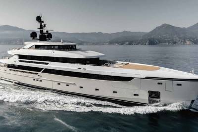 A sleek white yacht glides through the calm blue waters, with a mountainous coastline in the background.