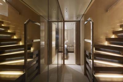 Illuminated wooden stairs ascend a luxurious hallway lined with mirrored closets reflecting a serene bedroom.