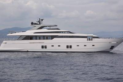 A sleek white yacht glides across the calm blue water with a coastal town visible in the distance.