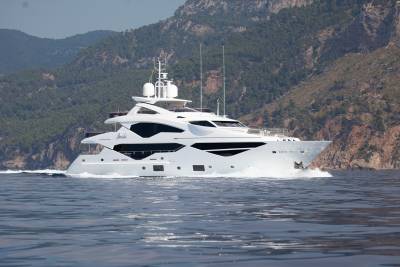 A sleek white yacht cruises through calm blue waters with a lush green mountainside in the background.