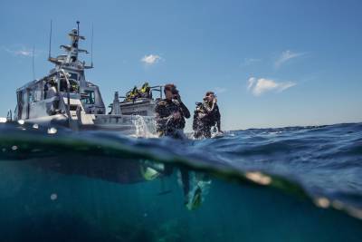 Divers in full gear prepare to enter the clear blue waters from a sleek boat under a bright sky.