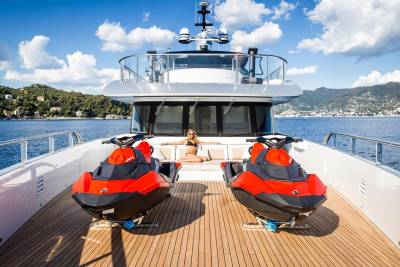 A woman relaxes on a luxury yacht's deck between two vibrant red jet skis, with a picturesque coastal town in the background.