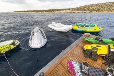 A collection of water toys, including a jet ski and inflatable tubes, are docked alongside a boat near a rocky coastline.