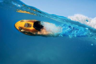 A person glides through the clear blue ocean water with a yellow underwater scooter, creating a trail of bubbles.
