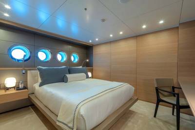 This luxurious yacht cabin features a comfortable bed, unique porthole windows with blue lighting, and elegant wood paneling.