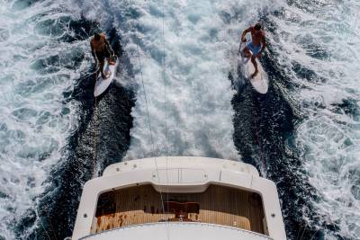 Two surfers ride the wake behind a luxury yacht, enjoying the thrill of the open water.