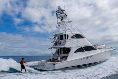 A surfer rides a wave behind a large white yacht under a cloudy blue sky.