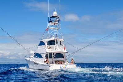 A luxurious sportfishing yacht cuts through the deep blue ocean under a bright sky with people fishing on deck.