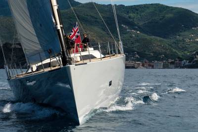 A sleek sailboat cuts through the azure waters with a Union Jack proudly displayed against a backdrop of verdant, villa-dotted hills.
