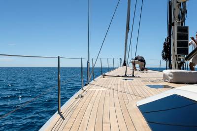 Two sailors work on the deck of a yacht sailing on a bright blue sea under a clear sky.