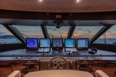 The yacht's advanced helm station offers panoramic views of the ocean at sunset, equipped with multiple navigation screens.