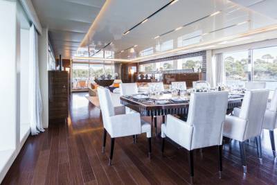 A luxurious yacht dining room features a polished wood floor, elegant white chairs, and a table set for a formal meal with a scenic view.