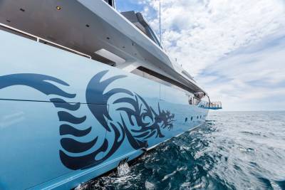 A striking blue yacht with a dramatic dragon decal glides through the shimmering ocean under a cloudy sky.