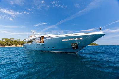 A sleek, blue yacht cruises through the sparkling blue waters of the Mediterranean Sea under a bright, cloudy sky.