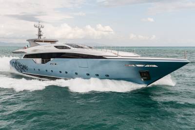 A sleek, light blue yacht with a dragon emblem cuts through the turquoise waters under a cloudy sky.