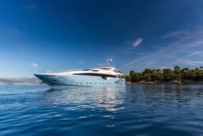 A sleek, light blue yacht with a dragon motif glides through calm, blue waters near a tree-lined coast.