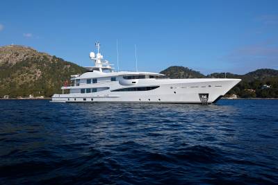A sleek white yacht glides across the deep blue sea, with lush green hills rising behind it under a clear sky.