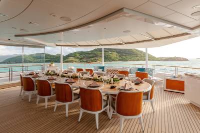 An elegant outdoor dining table is set for a feast on a yacht, overlooking a serene turquoise bay and lush green hills.