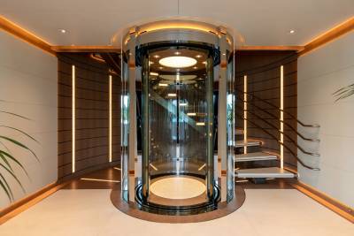 A luxurious glass elevator and curved staircase are illuminated by warm ambient lighting, creating a sophisticated interior.
