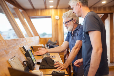 Two men consult a navigation screen on a wooden boat's bridge.