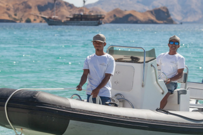 Two crew members in white t-shirts and caps stand ready on a rigid inflatable boat in the turquoise waters of Komodo National Park.
