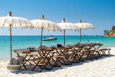 Elegant white umbrellas shade a long table set for a feast on a pristine beach beside turquoise waters.
