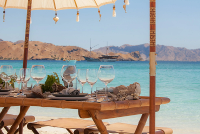 A beautifully set bamboo table awaits on a pristine beach, with crystal-clear turquoise waters and distant islands as the backdrop.