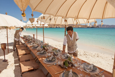 A long, beautifully set table awaits guests on a pristine beach, shaded by ornate umbrellas and overlooking turquoise waters.