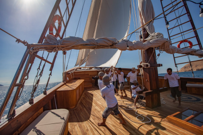 A crew works together to hoist the sails on a traditional wooden schooner under a bright blue sky.
