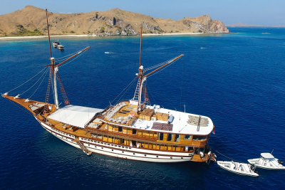 A majestic wooden schooner sails on a tranquil blue sea, anchored near a rugged, arid coastline.