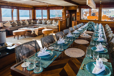 A long, elegant dining table is set for a feast aboard a luxurious wooden yacht, overlooking a serene body of water.