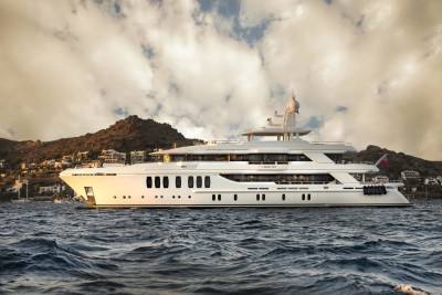 A luxurious white yacht named "Liquid Sky" cruises on choppy blue waters with a hillside village in the background under a dramatic sky.