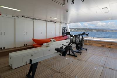 Two bright orange kayaks and a rowing machine are stored on the deck of a yacht overlooking a coastal town.