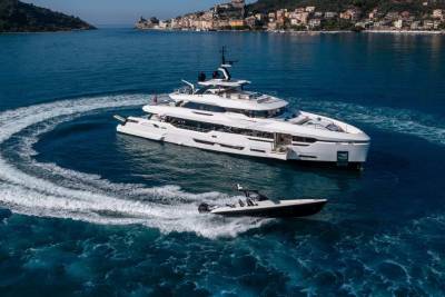 A large white yacht cruises through the deep blue sea, leaving a swirling wake, while a smaller boat speeds ahead.