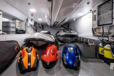Three colorful personal watercraft are neatly stored alongside dive tanks and a jet ski inside a boat's spacious garage.