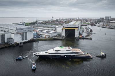 A massive yacht is guided out of a shipyard by two tugboats under a cloudy sky.