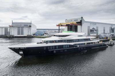 A sleek, dark-hulled superyacht glides from its shipyard under an overcast sky.
