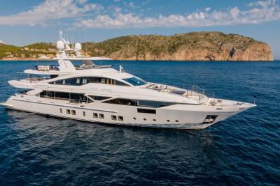 A luxurious white yacht glides through the deep blue waters off a rugged, tree-covered coastline under a bright blue sky.