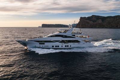 A sleek white yacht cuts through the dark blue sea, leaving a trail of white foam against a backdrop of rugged cliffs and a soft sunset sky.