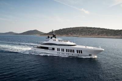 A sleek white superyacht cruises through the deep blue waters of a Mediterranean island chain under a clear sky.