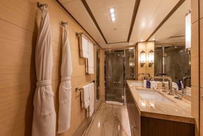 Luxurious white bathrobes hang ready in this opulent yacht bathroom with marble countertops and a glass-enclosed shower.