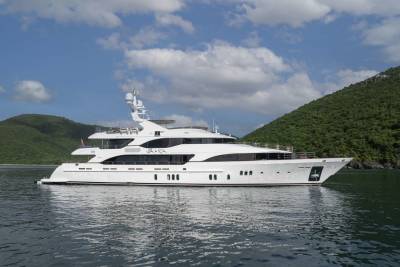 A luxurious white yacht glides through calm waters with lush green hills in the background under a cloudy blue sky.