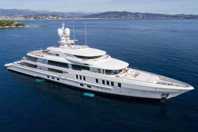 A luxurious white superyacht glides through the deep blue ocean, with a picturesque coastline in the background.