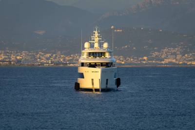 A luxurious white yacht glides across calm blue waters with a picturesque coastal city nestled at the base of distant mountains.