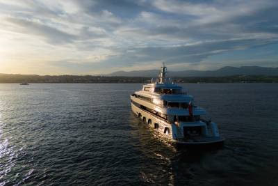 A luxurious superyacht glides across the tranquil sea at sunset, with a distant coastline bathed in golden light.