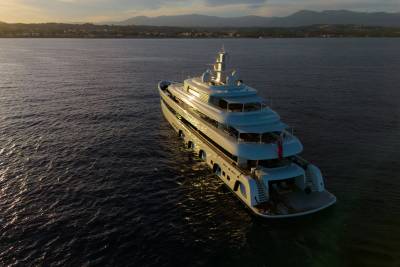 A luxurious white yacht glides across calm waters as the sun sets behind a distant coastal town.