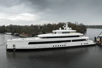A massive white superyacht glides through calm waters under a cloudy sky, flanked by trees and houseboats.
